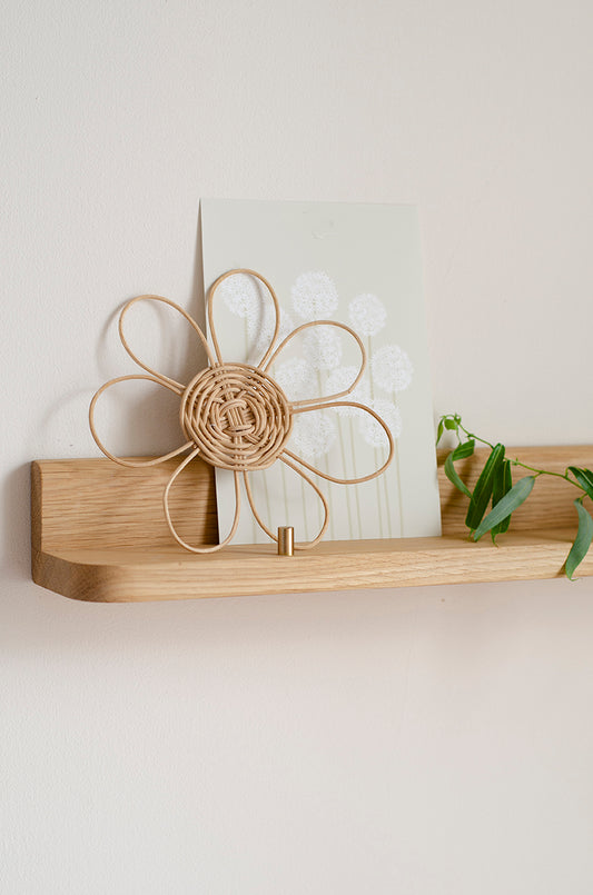 A light natural wood floating shelf mounted on a beige wall, holding a white card with a dandelion design and a small green plant sprig. A woven flower-shaped decorative object with a hook at the bottom is also resting on the shelf.