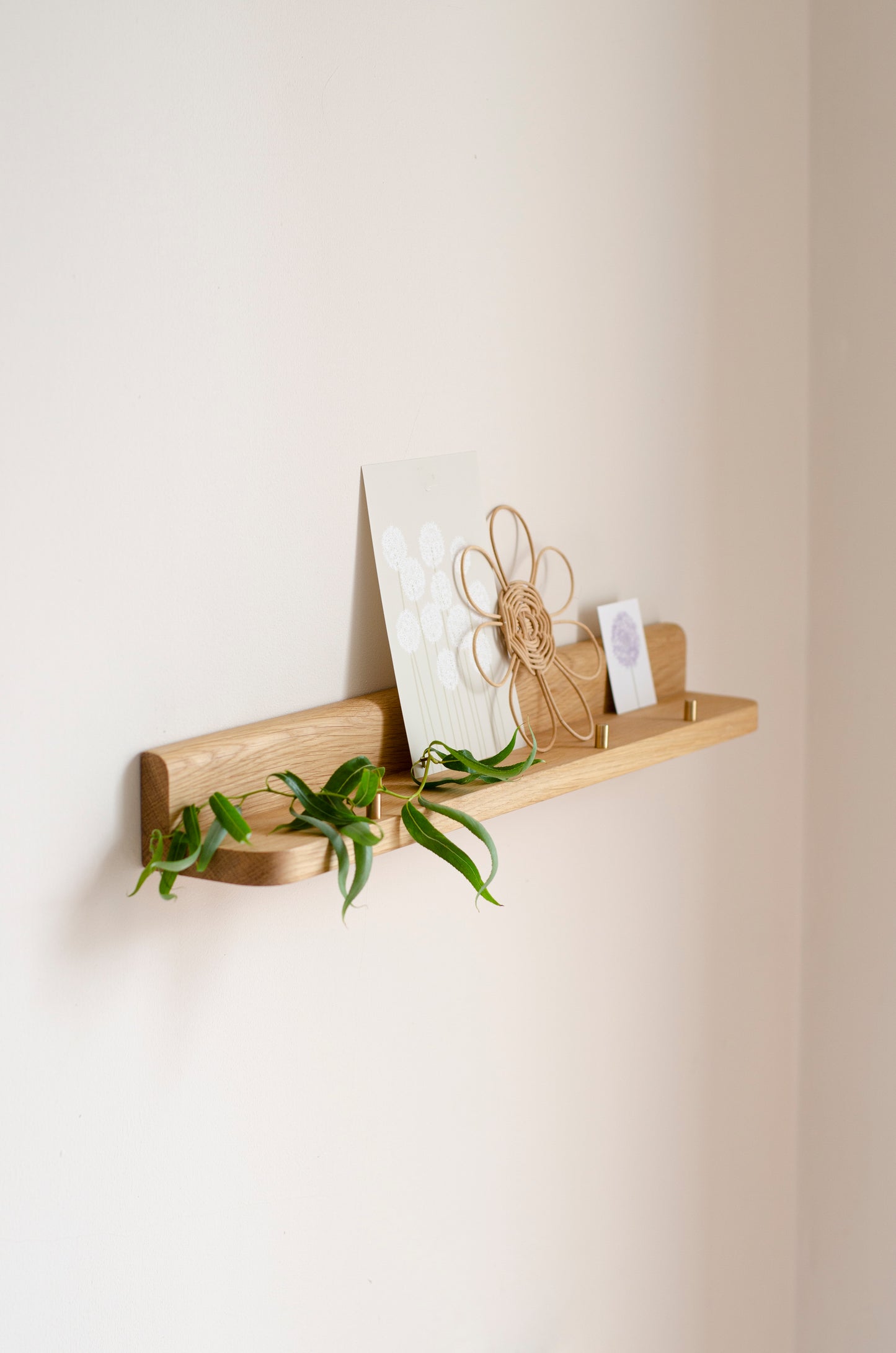A light natural wood floating wall shelf mounted on a warm beige wall, styled with a small green trailing plant, a card, and a decorative jute bow.
