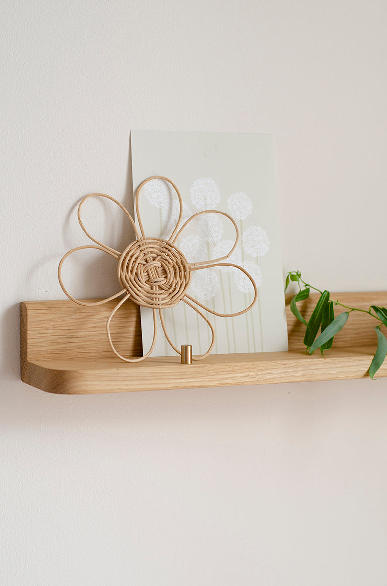 A light natural wood floating shelf mounted on a beige wall, holding a white card with a dandelion design and a small green plant sprig. A woven flower-shaped decorative object with a hook at the bottom is also resting on the shelf.