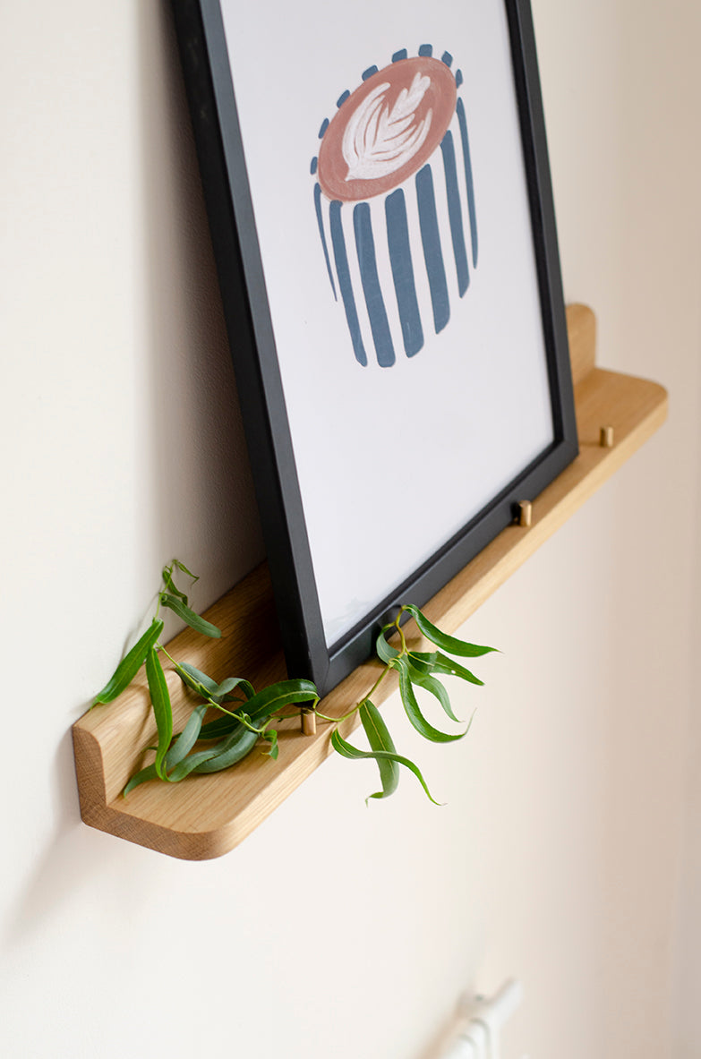 A framed wall art print with a minimalist coffee cup illustration in brown, beige, and navy blue, displayed on a light wood floating shelf with green willow leaves against a white wall.
